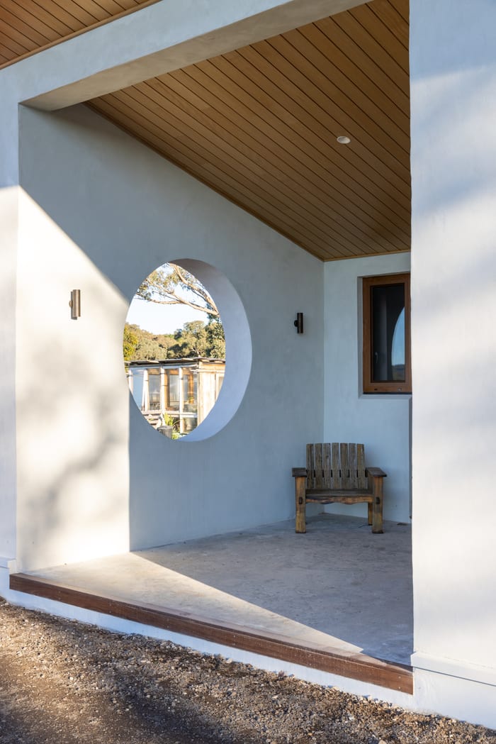 Round window punched through the rendered gable, seen from the porch under the deep timber-lined eave, with a timber bench below